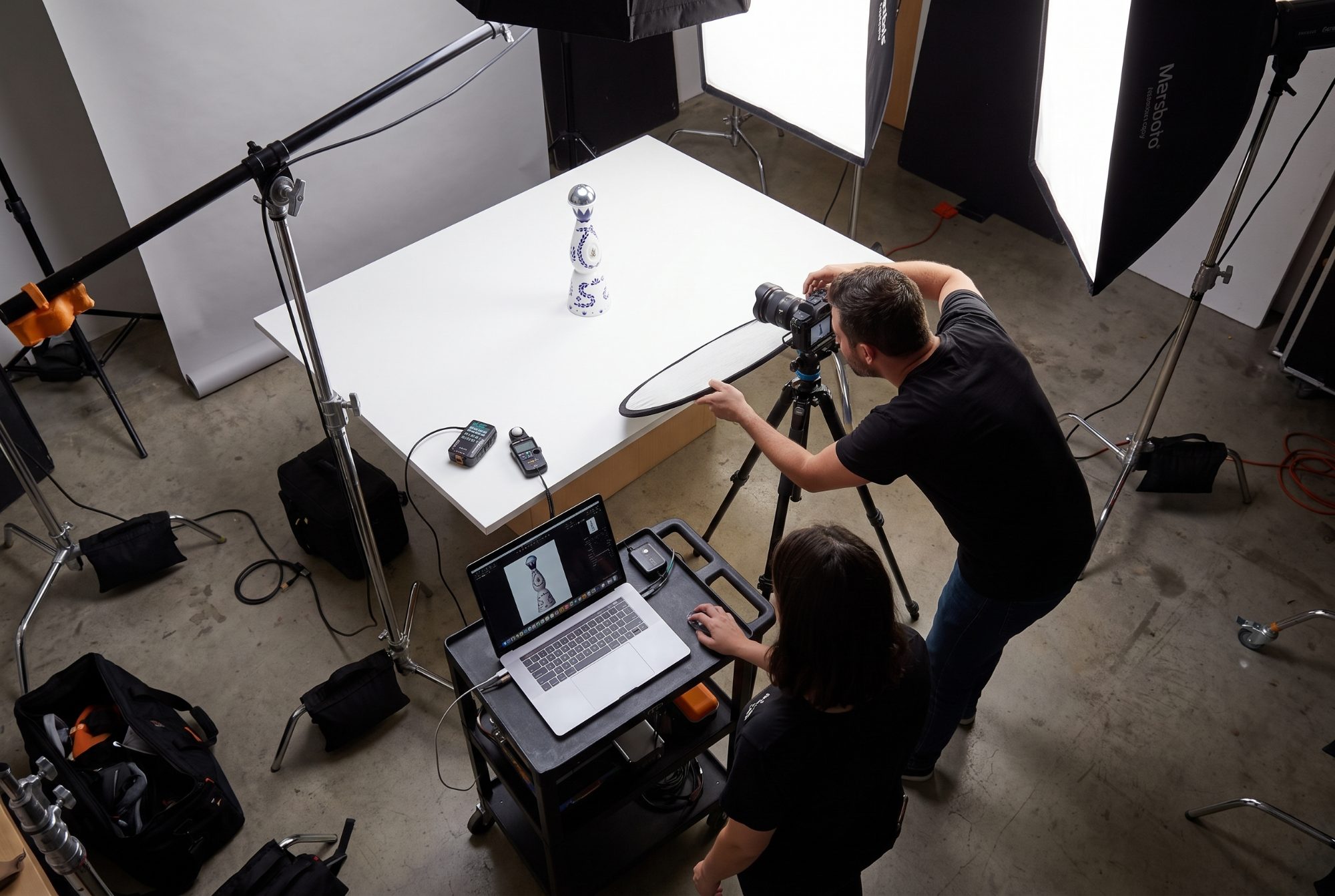 Overhead view of a commercial product photography studio — a bottle on a lit sweep, a tethered laptop showing the live capture, softboxes and stands around the set
